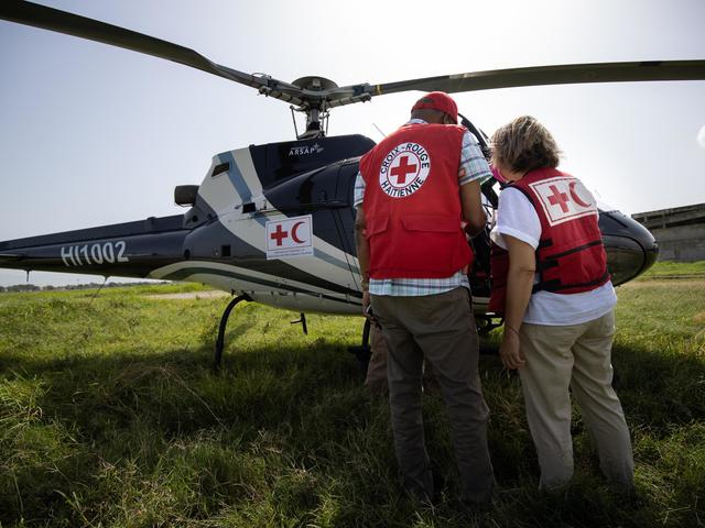 Two people standing in front of a helicopter with their back to the camera, both wear Red Cross / Red Crescent societies gear