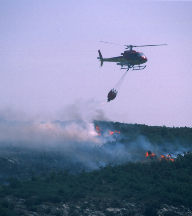 An Airbus-produced H125 helicopter deploys a bucket-type water dispersal system to support firefighting efforts.