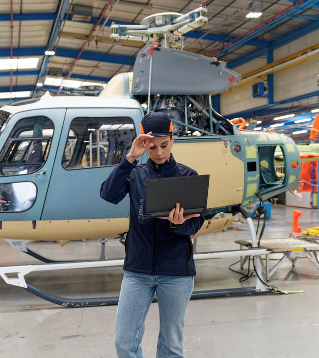 Woman wearing PPE stands in front of a helicopter looking at a laptop
